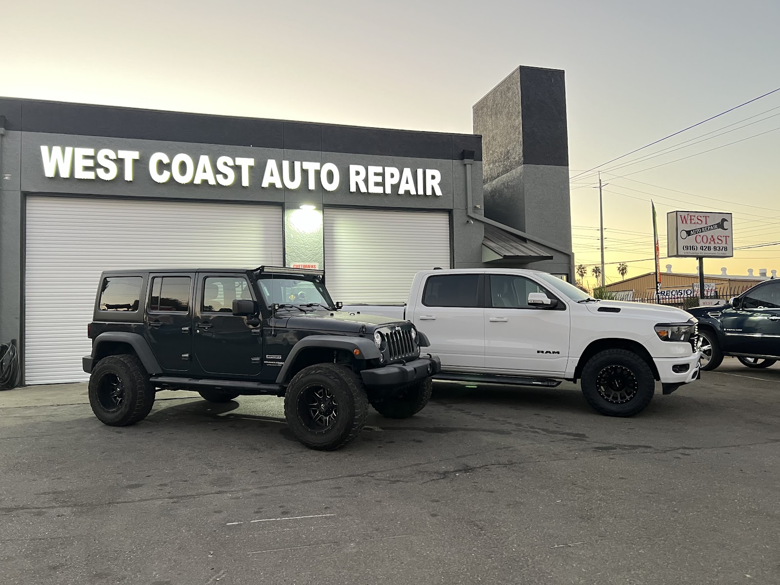 Two lifted trucks parked in front of the lit West Coast Auto Repair sign at dusk